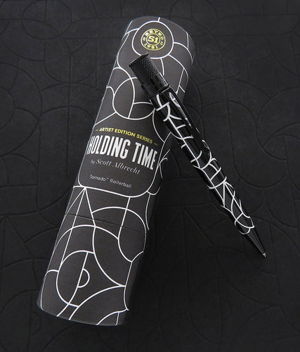 The pen rests on top of the presentation tube, in a dark background similar to the pen design.