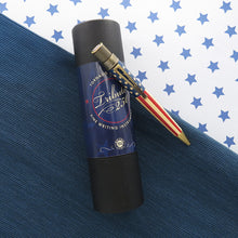 Load image into Gallery viewer, The rollerball pen leaning on the included gift tube. Set on a textured blue background split by a white background with stars. 
