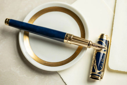 The Vitruvian Man Fountain Pen is set with the cap removed. The nib is leaning on the cap. The pen and cap are in a navy blue color, and the trims are golden. 