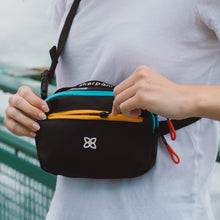 Load image into Gallery viewer, A person carrying the hip pack like a sling bag, while unzipping the front pocket. 

