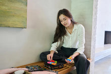Load image into Gallery viewer, Woman sitting on a wooden bench playing with the IQ Square Puzzle game.
