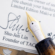 Load image into Gallery viewer, Close up to the fountain pen nib. It is two-toned, and engraved with the Taccia logo, and the text 18K 750 Japan. It is placed on top of the included certificate signed by Taccia founder: Shu-Jen Lin.
