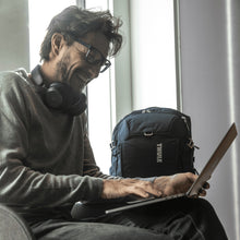 Load image into Gallery viewer, A young man sitting down and smiling while looking while viewing a laptop. The backpack is placed next to him. 
