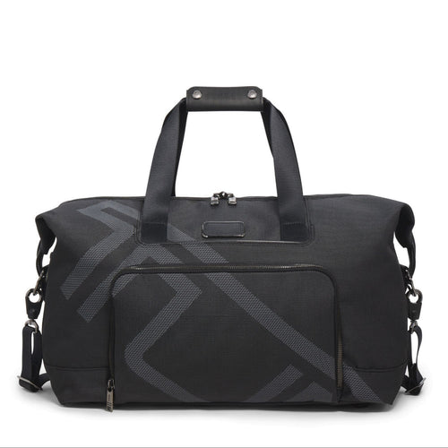 A black TUMI travel satchel with the logo printed across in light gray It features a top carry handle, detachable shoulder strap, and a front pocket. 