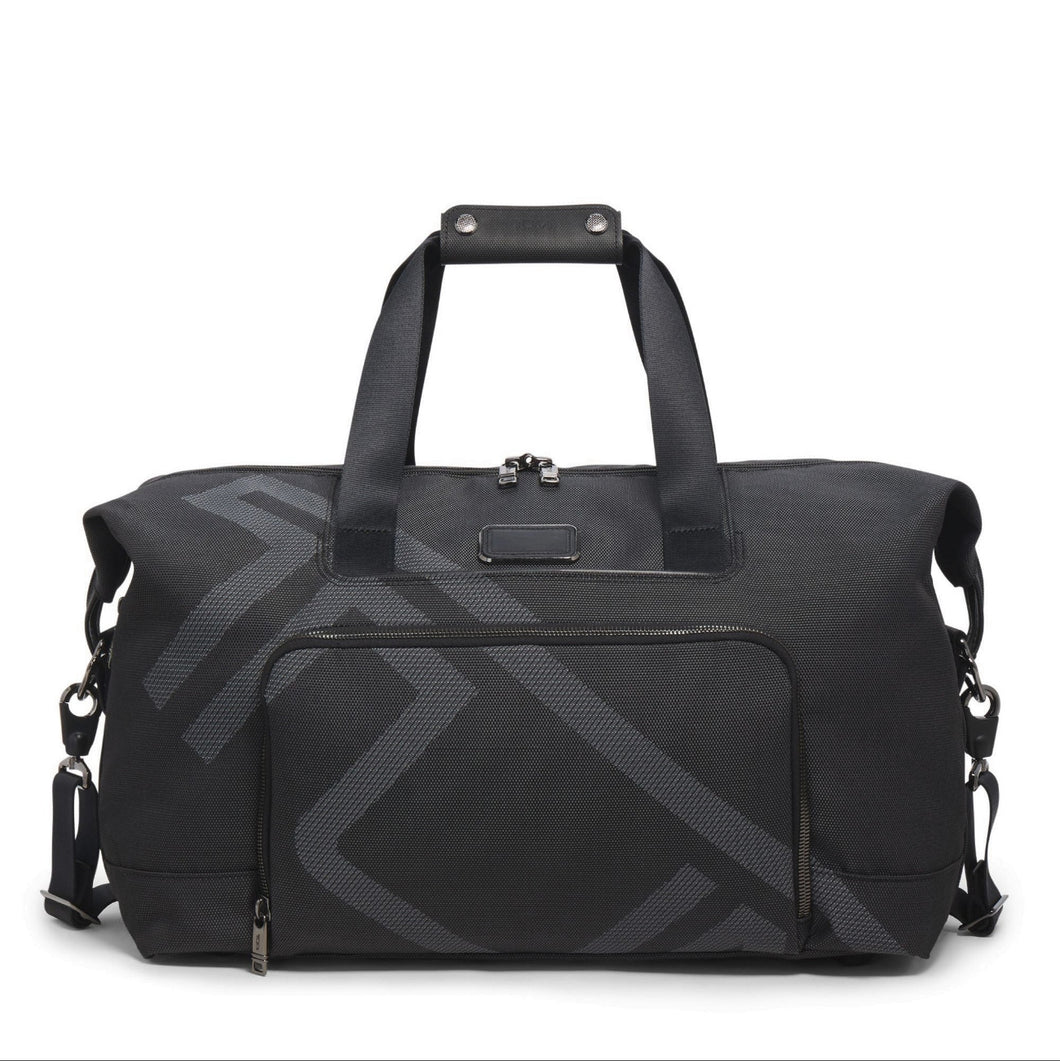 A black TUMI travel satchel with the logo printed across in light gray It features a top carry handle, detachable shoulder strap, and a front pocket. 