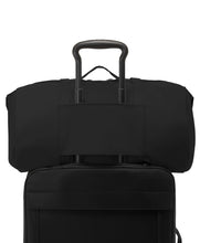 Load image into Gallery viewer, Back panel view of the duffle bag showing the attachment strap. A telescopic handle on a suitcase or trolley can slide through to hold the duffle in case.
