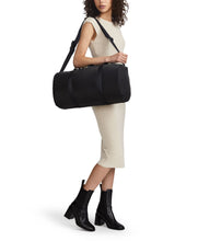 Load image into Gallery viewer, A young woman on a beige dress carrying the filled duffle bag on her shoulder with the adjustable strap.

