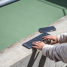 Load image into Gallery viewer, The keyboard is extended next to a cellphone on a concrete surface. A pair of hands type on the keys. 
