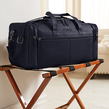 Load image into Gallery viewer, Angled view of the black leather duffle bag set on a foldable wooden stand in a hotel setting.

