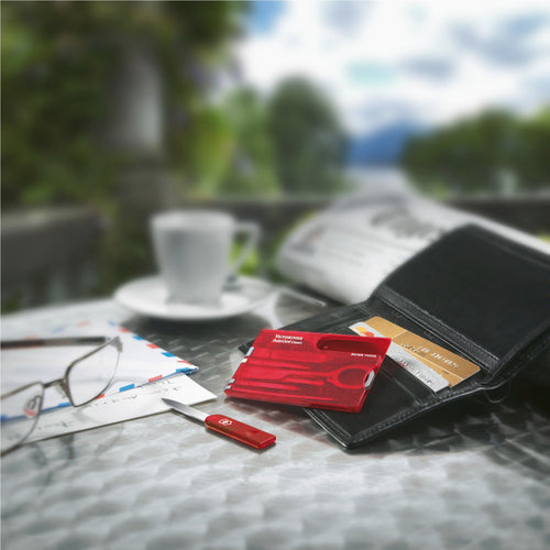 The tool swiss card placed on top of a wallet with the letter opener out. It is arranged on a table with a newspaper, a cup and saucer, and some glasses.