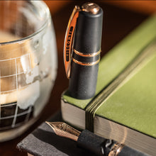 Load image into Gallery viewer, Uncapped fountain pen on a green notebook next to a glass container.
