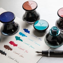 Load image into Gallery viewer, A navy blue, brown, turquoise, and red set of ink bottles arranged with a paper sampling of each, next to a fountain pen. 
