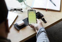 Load image into Gallery viewer, The charging station charging an earbuds case and smartwatch on top of a desk. A man holds his cellphone above the station.

