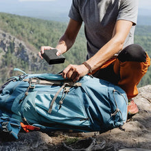 Load image into Gallery viewer, Close up to a person storing the folded charging station inside a hiking backpack. 
