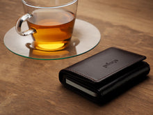 Load image into Gallery viewer, Angled view of the Pularis Boston Brown leather wallet closed on a brown wood surface. Some credit cards are visible from the side. A cup of tea is placed behind the wallet. 
