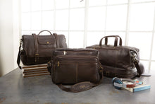 Load image into Gallery viewer, Two leather brown briefcases and a matching duffle bag arranged on a table by a window with a book and headphones. 
