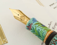 Load image into Gallery viewer, Close up to the fountain pen nib on top of the certificates. The grip section features a landscape of flowers and a blue bug. The nib features a stamp of the AP logo and the 14K-585 gold nib engraving.
