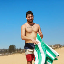 Load image into Gallery viewer, A young man wearing a swimming suit at the beach, holding the green and white towel.
