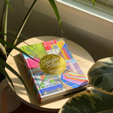 Load image into Gallery viewer, The Rainbow Atlas book arranged on a wooden table by a window, surrounded by plants.
