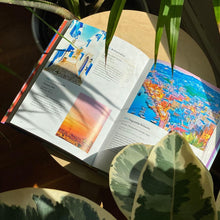 Load image into Gallery viewer, Interior view of the Rainbow Atlas book. Set on a wooden table by a window, surrounded by plants.
