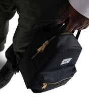 Load image into Gallery viewer, Angled close up to a a hand carrying the black Herschel nova Mini backpack by the top grip handle.
