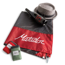 Load image into Gallery viewer, The picnic blanket arranged with a hat, wine bottle, and two folded blankets.
