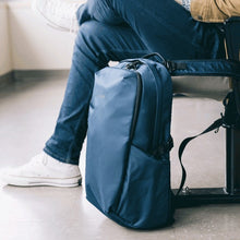 Load image into Gallery viewer, A young man sitting on a public area with the blue backpack attached to his seat.
