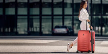 Load image into Gallery viewer, A young woman dressed in white, crossing the street as she pulls her red Victorinox suitcase. 
