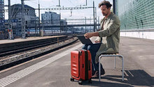 Load image into Gallery viewer, A man sitting on a bench by a train station with a Red Victorinox suitcase. 
