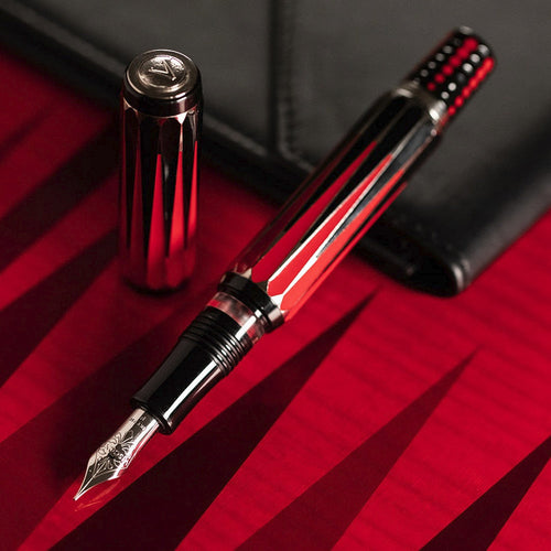 The Visconti Backgammon Fountain Pen with the cap set behind it. It is laid on a red background and leaning on a black leather letter pad.